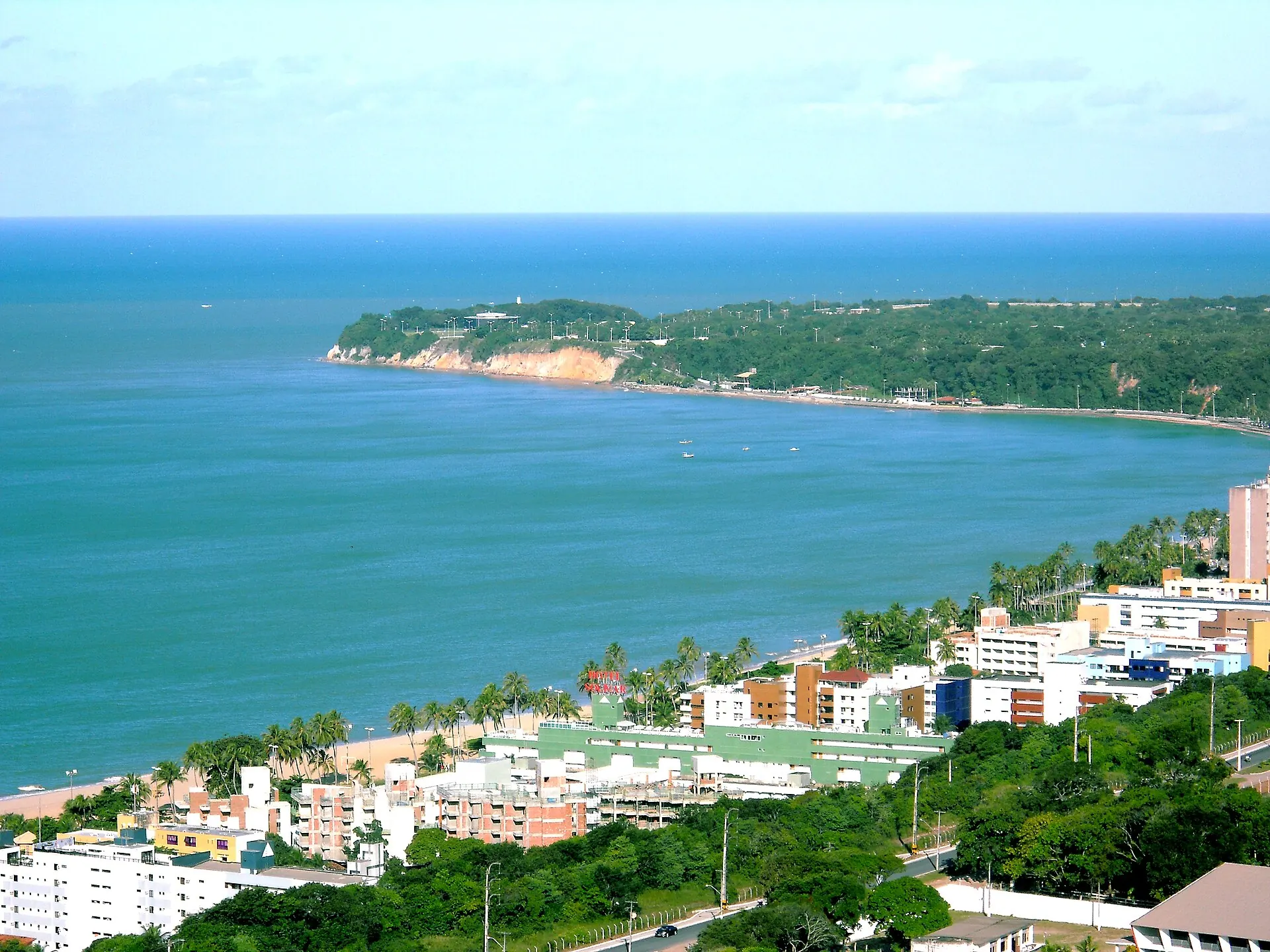 Praia do Cabo Branco vista do alto, com o mar esverdeado e a ponta do Cabo Branco coberta de mata ao fundo