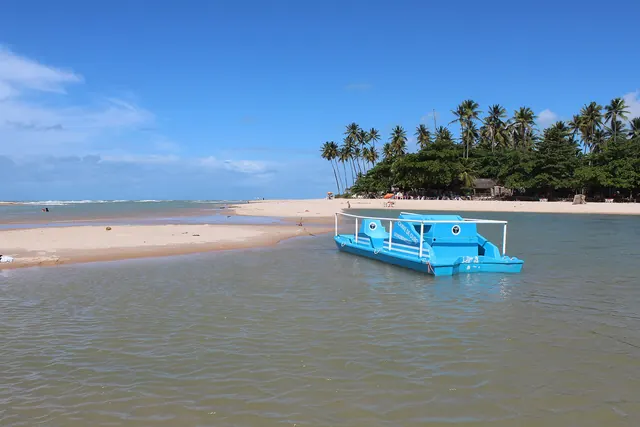 Foz do rio Gramame com água rasa, um barco azul de passeio, banco de areia e coqueiros na outra margem