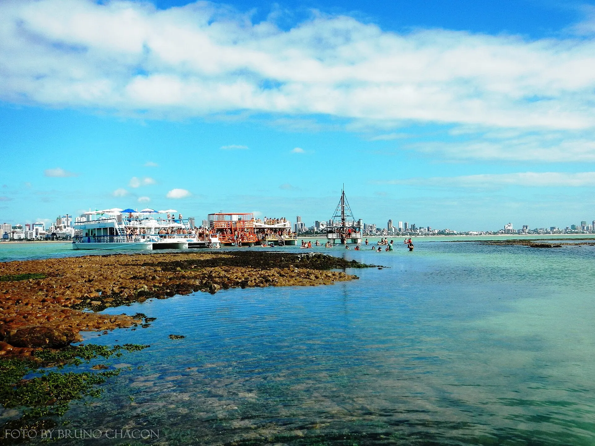 Recife de Picãozinho descoberto na maré baixa, com poças de água clara, barcos de passeio ao longe e os prédios da orla de João Pessoa no horizonte