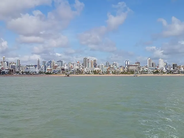 Orla de João Pessoa vista do mar, com a linha de prédios na beira da praia, mar verde e céu azul com nuvens