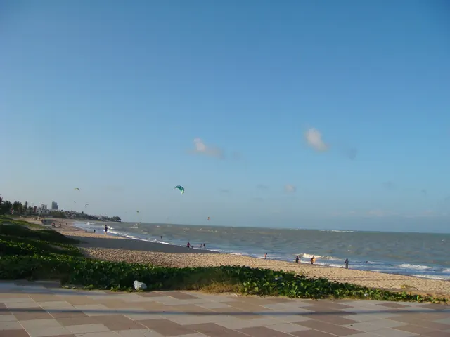 Praia do Bessa vista do calçadão, com vegetação de restinga, areia larga, mar calmo e pipas de kitesurf no céu