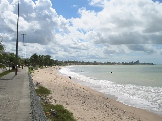 Praia de Manaíra com o calçadão à esquerda, faixa de areia, ondas fracas e a Praia do Bessa ao fundo sob nuvens