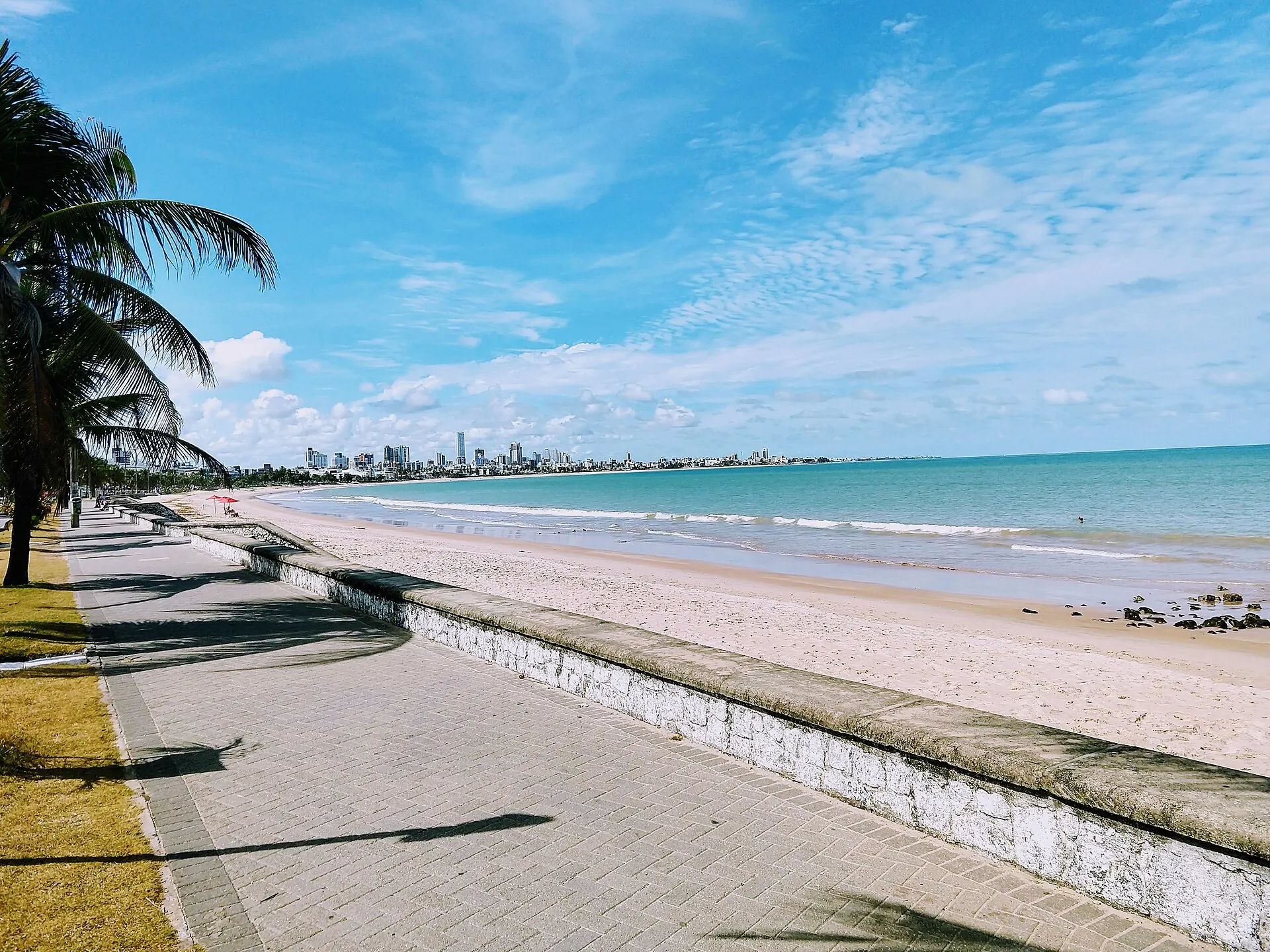 Calçadão à beira da praia na orla de João Pessoa, com areia clara, mar azul e prédios no horizonte